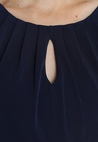 Marinblå klänning med plisserad halsringning och en nyckelhålsdetalj. Slät, texturerad tyg. Nära vy av övre området.