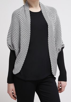 Kobieta ubrana w czarny top z długim rękawem i spodnie z białym otwartym kardiganem w wzór sześciokątów na przodzie.
