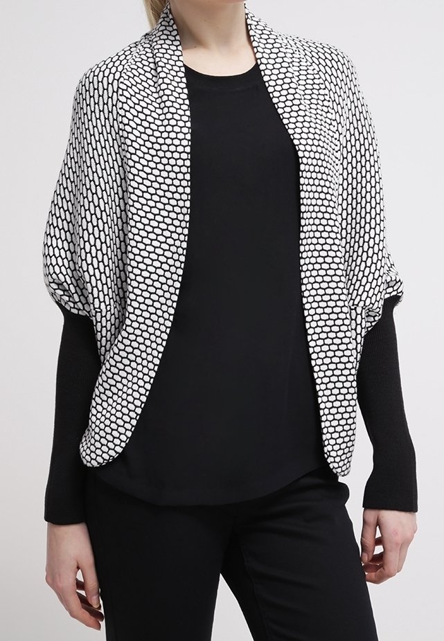 Femme portant un haut noir à manches longues et un pantalon avec un cardigan blanc à motif hexagonal ouvert sur le devant.
