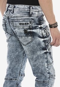 Blaue Denim-Cargo-Jeans mit einem verblassten, abgetragenen Muster. Verfügt über mehrere Taschen und dekorative Nähte. Akzentuiert mit schwarzem Branding.
