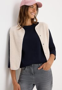 Leende kvinna med rosa keps, krämfärgad knäppt fleeceväst över en marinblå långärmad tröja och grå jeans, med en hand i fickan, lutande mot en vit vägg.