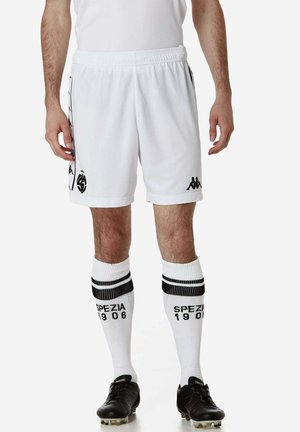 Pantaloni corti da calcio bianchi con logo e dettagli neri, abbinati a calze bianche al ginocchio con strisce nere e testo "SPEZIA 1906". Scarpe nere.