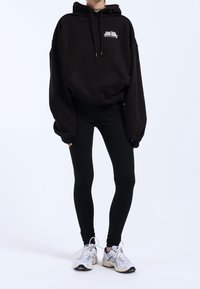 Černá oversized mikina s kapucí a přední kapsou, s bílým logem, v kombinaci s černými legínami a vícebarevnými sportovními botami. Jednoduchý a moderní design.