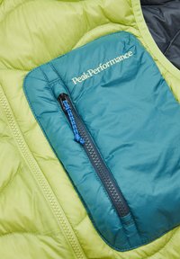 Grüne gesteppte Jacke mit einer türkisfarbenen Reißverschlusstasche. Die Tasche hat einen blauen Zipper und das "PeakPerformance"-Logo in Weiß.