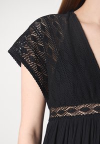 Μαύρο φόρεμα με περίπλοκες δαντελένιες λεπτομέρειες στις μανίκες και τη μέση, σε ρευστό ύφασμα και σχέδιο V-λαιμού.