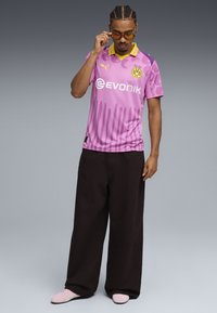 Camiseta de fútbol rosa con acentos en negro y amarillo, que presenta un sutil patrón de rayas, combinada con pantalones sueltos negros y zapatos rosa claro.
