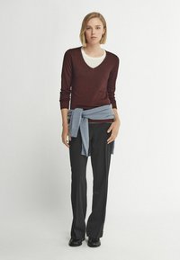 Burgunderfarbener V-Ausschnitt-Pullover, über einem hellen Oberteil getragen, kombiniert mit schwarzen Hosen und einem hellblauen Pullover, der um die Taille gebunden ist. Schwarze Schuhe getragen.