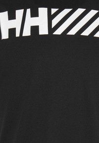 Prenda negra con tejido texturizado, que presenta grandes letras blancas y rayas diagonales. Centrada en elementos de diseño deportivos.