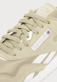Κοντινή λήψη μπεζ αθλητικού παπουτσιού Reebok που δείχνει υφή σουέντ και πλέγματος, μπεζ κορδόνια, λευκές ρίγες και το λογότυπο Reebok με τη σημαία του Ηνωμένου Βασιλείου στην πλευρική επιφάνεια.