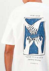 T-shirt en coton blanc avec un graphisme bleu représentant des mains entrelacées et un texte en dessous. Coupe décontractée avec des manches courtes et un col rond.