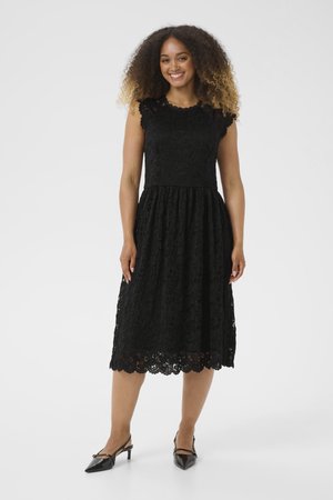 Black lace dress with sleeveless design, scalloped hem, and flared skirt. Paired with black pointed-toe heels, showcasing intricate details.