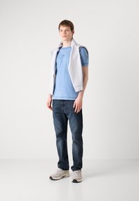 T-shirt de manga curta azul claro com gola redonda e logotipo, combinado com jeans azul escuro e ténis bege. Destacado com um suéter cinza colocado sobre os ombros.