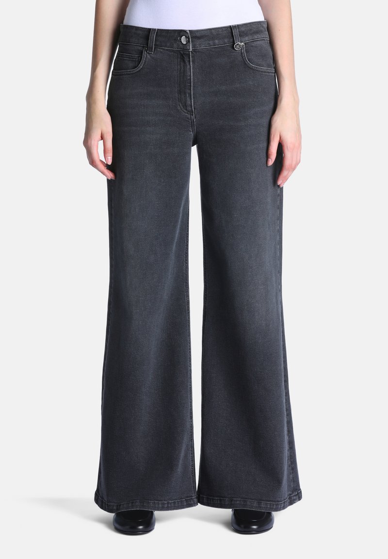 Jeans noirs à jambes larges portés par une personne debout, associés à des chaussures noires et un haut blanc visible à la taille.