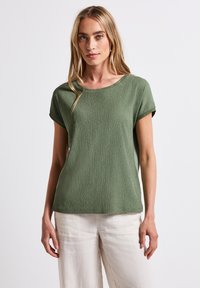 Olivgrön topp med korta ärmar, texturerat mönster och rund halsringning, tillsammans med ljusa, lediga byxor.