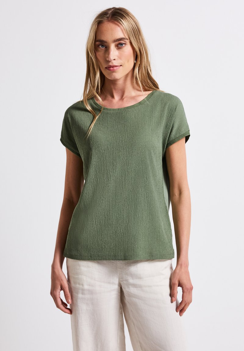 Olivgrön topp med korta ärmar, texturerat mönster och rund halsringning, tillsammans med ljusa, lediga byxor.