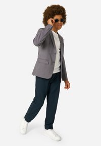Grauer Blazer mit Reverskragen, kombiniert mit einem weißen T-Shirt, dunkelblauer Hose und weißen Sneakern. Accessoires umfassen runde Sonnenbrillen.