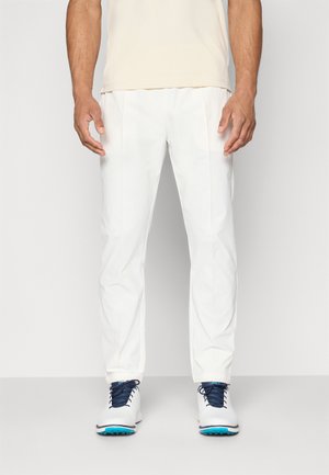 Weiße Golfhose mit geradem Schnitt, aus glattem Material und mit dezenten Bügelfalten, kombiniert mit weißen und blauen Golfschuhen.
