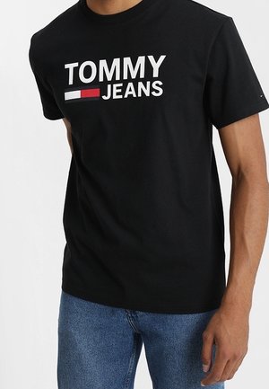 Mann trägt ein schwarzes T-Shirt mit dem Aufdruck "TOMMY JEANS" und einem rot-weiß-blauen Logo, kombiniert mit blauen Jeans.