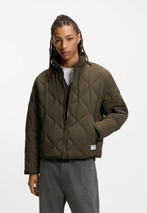 Ung person med langt fletningshår iført olivengrøn quiltet jakke med lynlås og grå bukser, stående foran en ensfarvet hvid baggrund.