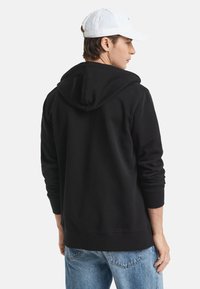 Schwarzer Kapuzenpullover mit Rippbündchen und Saum. Dazu werden hellblaue Jeans getragen. Auf dem Kopf des Models sitzt eine weiße Baseballkappe.