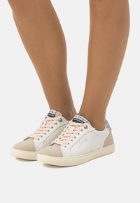 Zapatillas blancas y beige con cordones rosas, un upper de cuero suave y una puntera texturizada. Presentan una punta redonda y un logotipo en la lengüeta.