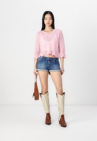Rózsaszín horgolt crop top V-nyakkal, farmer sort és bézs cowboy csizmát viselve, barna krokodilbőr hatású táskát tartva.