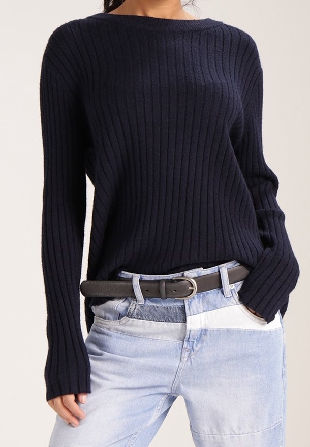 Personne portant un pull côtelé bleu marine à manches longues, rentré dans un jean patchwork clair avec une ceinture noire.