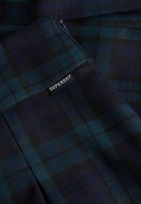 Tela de cuadros con tonos azul oscuro y verde, que presenta una etiqueta de Superdry y una textura suave. El diseño incluye líneas entrecruzadas.