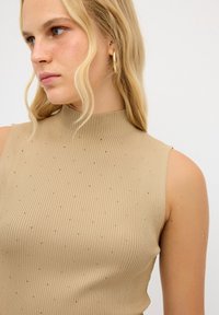 Top smanicato beige a costine con collo alto, tessuto texturizzato e piccoli dettagli dorati, caratterizzato da un design aderente.