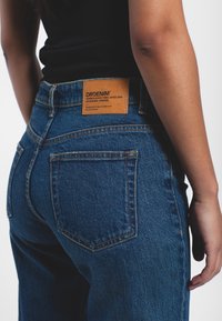 Blue denim jeans with a high waist, featuring a tan leather tag on the back waistband and subtle stitching detail on pockets.