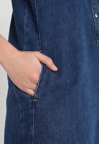 Denim-mekko, jossa on syvänsininen väri, nappisulku ja sivutaskut, joissa on keltaiset ompelukset. Texturoitu kangas, jossa on näkyvä puu.