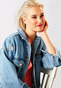 Giacca in denim di colore blu sbiadito, con dettagli usurati e bottoni argentati. Il modello ha capelli corti biondi e sieda su una sedia bianca.