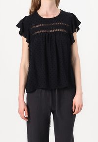 Femme portant un blouse noire à manches courtes avec un panneau transparent à pois et des manches volantées, associée à un pantalon noir texturé ample.