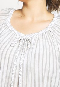 Blusa ligera de rayas con cuello redondo, cierre con lazo trenzado y tela texturizada en tonos suaves de blanco y gris.