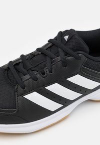 Black athletic shoe with a textured synthetic upper, featuring three white stripes and black laces. Rubber outsole with a gum accent.