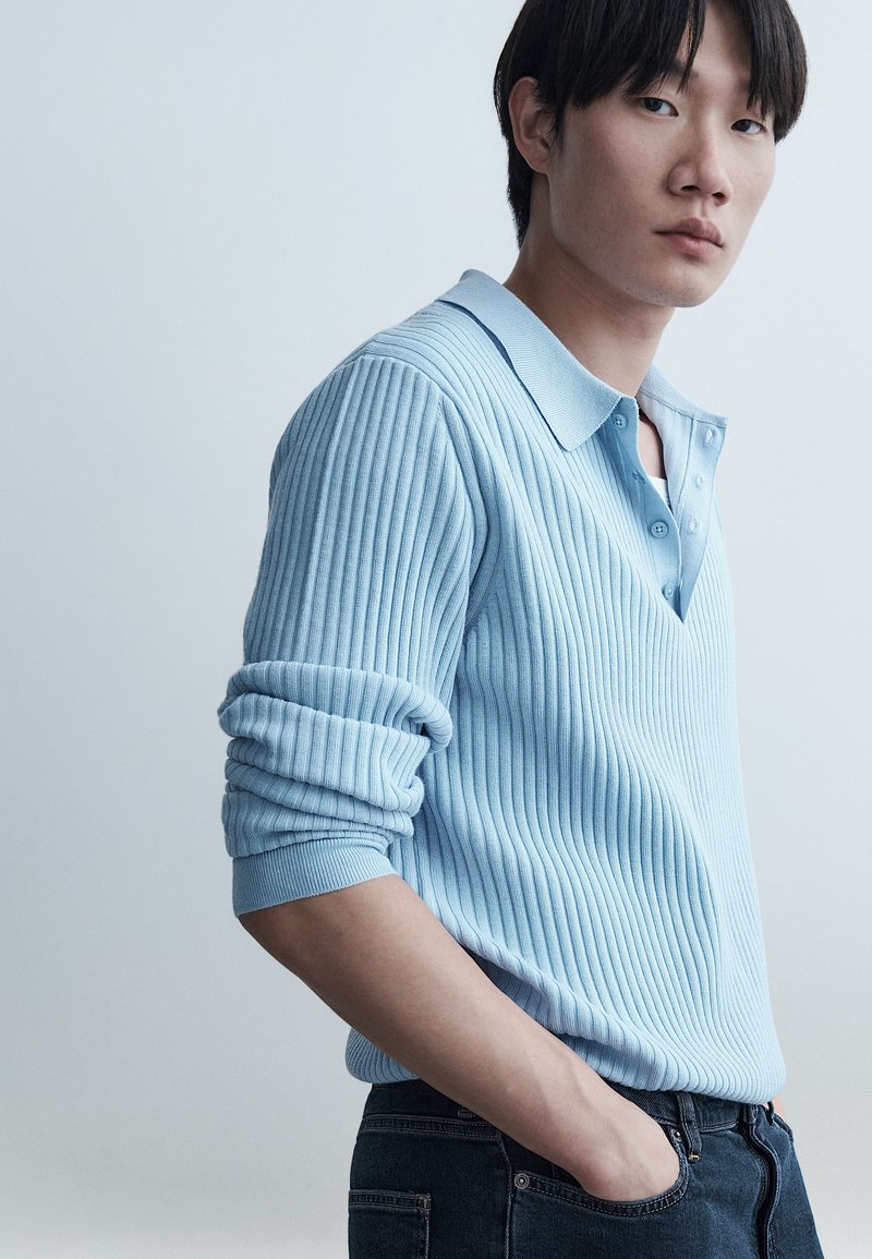 Jeune homme aux cheveux noirs portant un pull côtelé bleu clair à manches retroussées et un jean foncé, debout avec une main dans la poche.