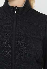 Doudoune sans manches noire avec un col haut, présentant un motif texturé et une fermeture éclair à l'avant, associée à un tissu noir lisse sur les côtés.