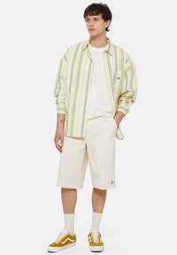 Chemise à rayures, jaune clair et verte, t-shirt blanc, shorts crème et baskets moutarde avec accents blancs et chaussettes mi-mollet.