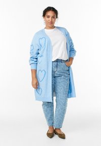 Svetlo modrý kardigán s motívmi srdca. Mäkká pletená tkanina, otvorený predný dizajn, v kontraste so svetlo modrými vysoko pásovými džínsami. Hnedé baleríny.