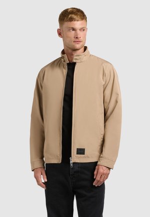 Muž oblečený v béžovej bunde na zips s stojacím golierom, pod ktorou má čierne tričko a čierne nohavice, mierne sa pozerá do strany.