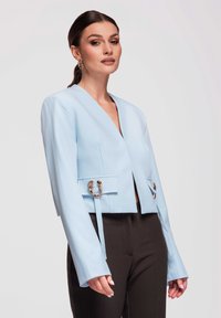Femme portant un blazer court bleu clair avec des détails de boucle dorée et un pantalon noir taille haute, debout devant un fond uni.