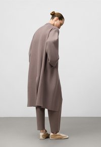 Una donna con un chignon indossa un cappotto oversize color taupe e pantaloni abbinati, in posizione laterale, con le mani nelle tasche, sneakers beige visibili su un pavimento grigio.