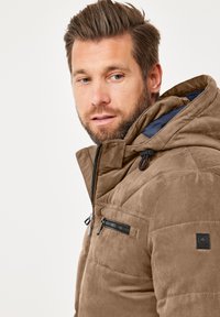 Brown quilted jacket with a hood, featuring a zippered chest pocket and textured fabric. Black accent logo on the sleeve.