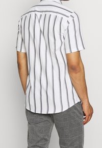 Uomo vestito con una camicia bianca a maniche corte con strisce verticali nere e pantaloni a quadri grigi, mostrato di spalle su uno sfondo semplice.