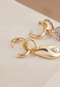 Zlate kovinske uhane s ukrivljenimi, skulpturalnimi oblikami in majhnim bisernim dodatkom, prikazani na teksturirani bež površini.