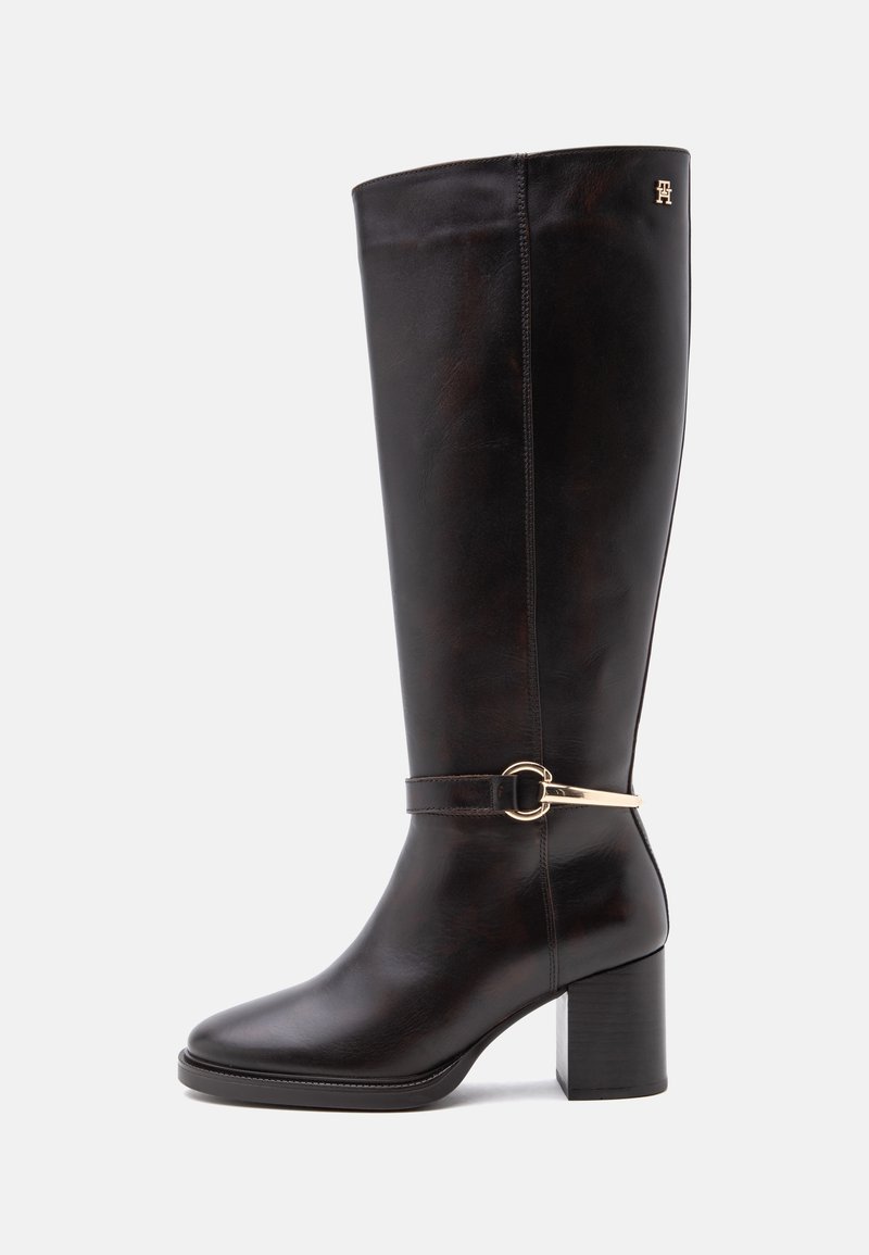 Botte en cuir noire montant jusqu'au genou avec talon bloc et détail de sangle à boucle dorée près de la cheville sur un fond clair uni.