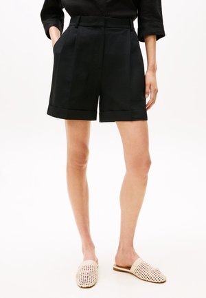 Kvinde iført sorte skræddersyede shorts med høj talje og opsmøgede kanter, en sort top og beige vævede slip-on sko, stående med den ene hånd i lommen.