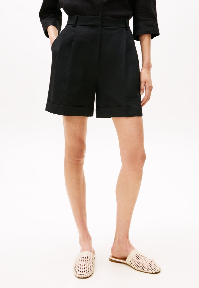 Femme portant un short noir taille haute sur mesure avec ourlets retournés, un haut noir et des chaussures beiges tissées à enfiler, debout avec une main dans la poche.