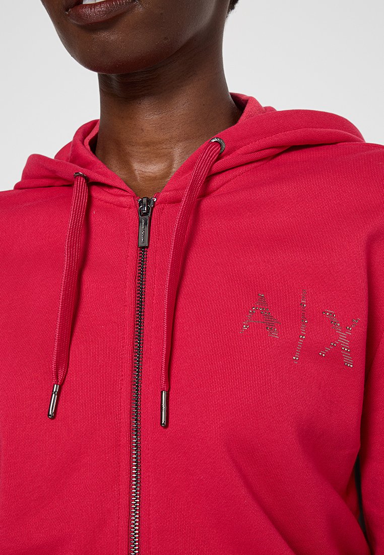 Persona che indossa una felpa rossa con zip e cappuccio, decorata con un piccolo logo "A|X" sul petto.