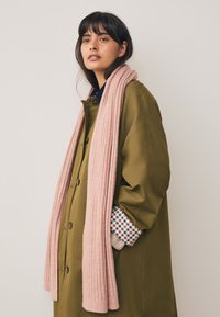 Olivengroene, knoopsluitende jas met een ontspannen pasvorm, gecombineerd met een geribde roze sjaal. De jas heeft een geruit stof zichtbaar aan de manchetten.